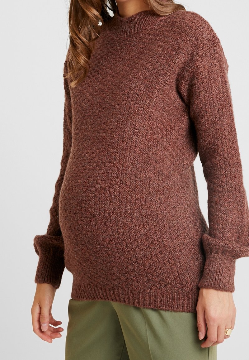 Těhotná žena v hnědém pleteném svetru s texturou a olivově zelených kalhotách, ukazující svůj těhotenský břicho.