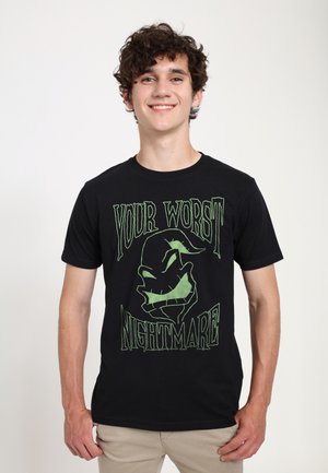 Jeune homme aux cheveux bouclés souriant, portant un t-shirt noir avec un texte graphique vert "Votre pire cauchemar" et une illustration stylisée d'un visage.