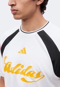 Άνδρας που φοράει λευκή και μαύρη μπλούζα Adidas με κίτρινο λογότυπο και επιγραφή Adidas, εμφανίζεται από τους ώμους και πάνω, κοιτάζει αριστερά.