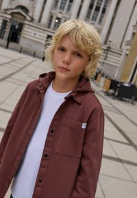 Veste boutonnée bordeaux avec capuche, poche avant et fermetures à pression noires, superposée sur un T-shirt blanc. Tissu texturé, design décontracté.