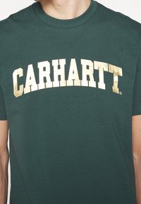 Zielona bawełniana koszulka z krótkimi rękawami z dużym, zakrzywionym, metalicznym złotym napisem "CARHARTT" na piersi.