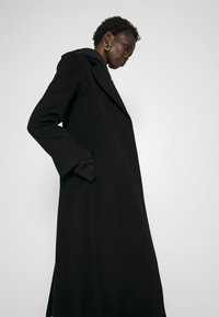 Langer schwarzer Mantel mit Kapuze, der eine taillierte Silhouette, breite Revers und versteckte Taschen aufweist. Glatte Textur mit einem minimalistischen Design.