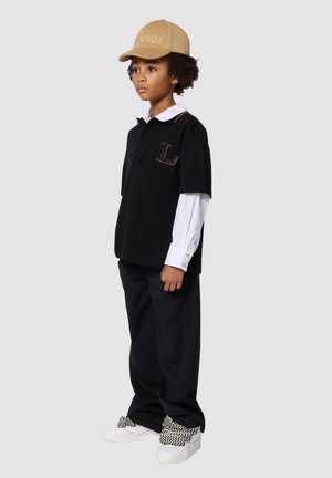 Polo noir à manches courtes avec une chemise blanche à manches longues en dessous, un pantalon noir et une casquette beige ; présente des baskets blanches avec des lacets à motifs.