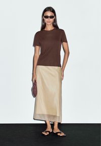 Brown cotton t-shirt, beige satin maxi skirt, black flat sandals. Model holds a textured maroon clutch. Simple design, neutral colors.