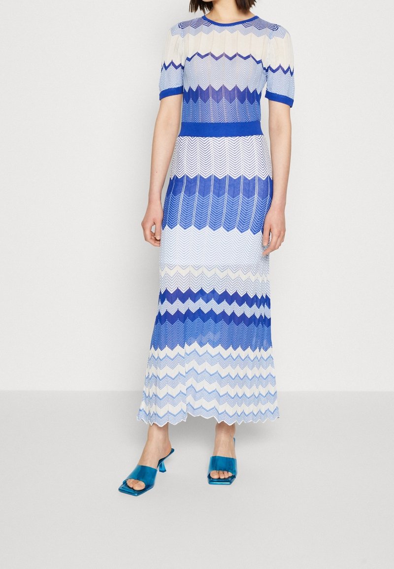 Robe maxi avec un motif en zigzag dans différentes nuances de bleu et blanc, fabriquée dans un matériau léger, associée à des sandales à talons bleues.