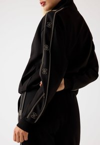 Giacca sportiva nera con strisce accentate dorate e dettagli con logo sulle maniche. Presenta un collo alto e tasche con zip per la funzionalità.