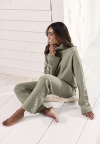 Olivgrünes Strick-Loungewear-Set mit Rollkragen und lockerem Schnitt. Die Hose hat ein seitliches Logo-Detail. Weicher Stoff, minimalistisches Design.