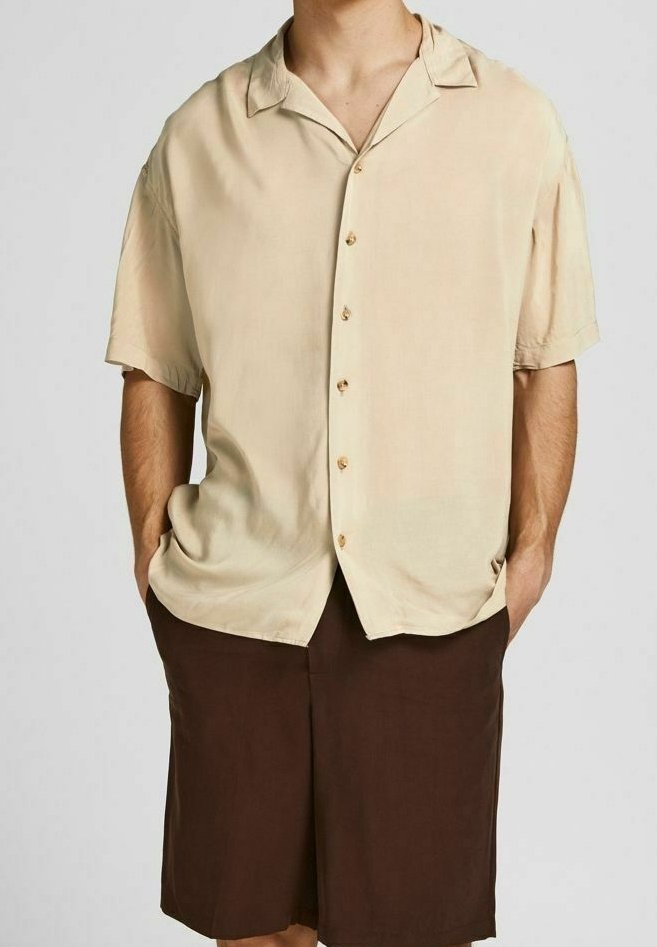 Homme portant une chemise beige à manches courtes avec col et un short marron longueur genoux, mains dans les poches.