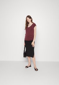 Top burgundy de mangas curtas e decote em V, combinado com uma saia lápis preta com textura. O modelo segura uma bolsa preta e usa sandálias deslizantes pretas.