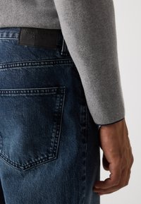 Close-up of a person’s hand resting by their side, wearing dark blue denim jeans and a grey long-sleeve top.