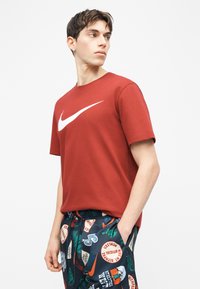 Νέος άνδρας με κόκκινο μπλουζάκι Nike και αθλητικό σορτς με σχέδια, με τα χέρια στις τσέπες, κοιτάζει προς το πλάι.