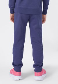 Persoon in een marineblauwe sweatpants en bijpassende sweatshirt, staand, met roze sportschoenen op een witte achtergrond.