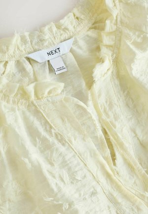 Hellgelbes Oberteil aus strukturiertem Stoff mit gerafftem Halsausschnitt und Vorderöffnung. Das Etikett trägt die Aufschrift "NEXT", hergestellt in Kambodscha.