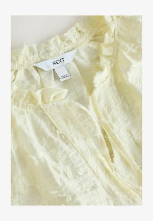 Hellgelbes Oberteil aus strukturiertem Stoff mit gerafftem Halsausschnitt und Vorderöffnung. Das Etikett trägt die Aufschrift "NEXT", hergestellt in Kambodscha.