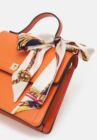 Sac à main en cuir orange avec bordure noire, doté d'un fermoir doré et d'un foulard en soie à motif attaché en accent. Forme rectangulaire compacte.