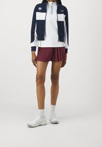 Marinblå och vit zip-up jacka med färgblockdesign, ihop med plisserade bourgogne-shorts och vita träningsskor. Strumpor är vita.