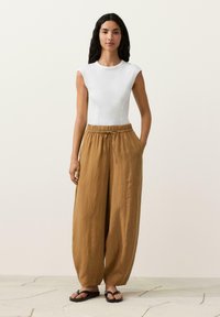 Hosszú sötét hajú nő, fehér ujjatlan felsőt, laza barna zsinóros nadrágot és fekete papucsot visel, csempézett padlón áll.