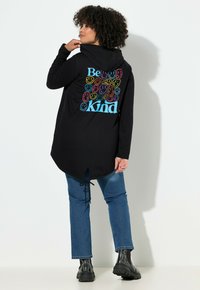 Persona con cabello rizado que lleva un sudadera con capucha negra con un diseño colorido de "Sé amable" y una cara sonriente, jeans azules y botas negras, de pie de lado.