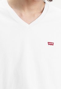 T-shirt blanc à col en V en coton ; présente un petit logo rouge Levi's sur la poitrine gauche. Texture lisse avec une coupe classique.