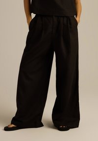 Czarne spodnie o szerokich nogawkach, z plisowanym przodem, dwoma bocznymi kieszeniami i gładką teksturą, zestawione z płaskimi sandałami.