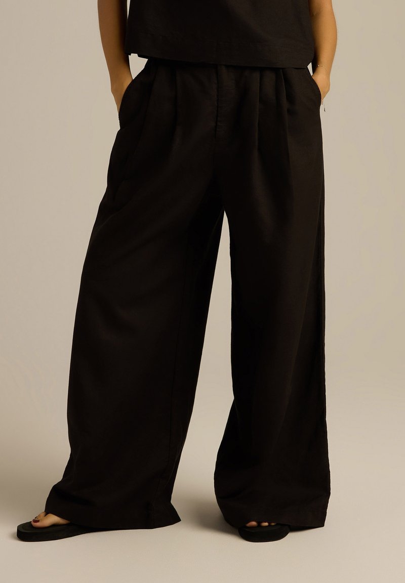 Czarne spodnie o szerokich nogawkach, z plisowanym przodem, dwoma bocznymi kieszeniami i gładką teksturą, zestawione z płaskimi sandałami.