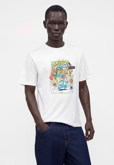 Άνδρας που φοράει λευκό T-shirt με έγχρωμο καρτούν γραφικό και το κείμενο «Μην παίρνεις τη ζωή πολύ σοβαρά», συνδυασμένο με σκούρα μπλε τζιν.