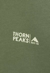 Tessuto verde con una superficie liscia che presenta il testo bianco "THORN PEAKS" e un logo di montagna, insieme a "ROUTE 189" in un design semplice.