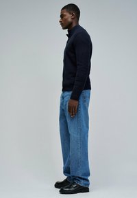 Jersey de punto azul marino con cuello alto, combinado con jeans de pierna recta azul claro y mocasines negros, visto de lado.