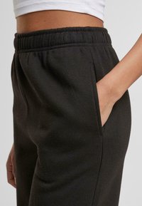 Personne portant un pantalon de jogging noir à taille élastique avec la main dans la poche latérale et un crop top blanc visible au niveau de la taille.