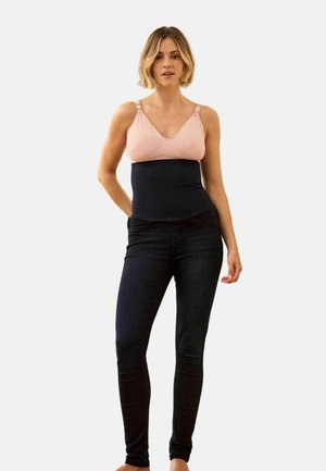 Woman wearing a pink nursing bra and black high-waisted maternity jeans, standing against a plain light background.