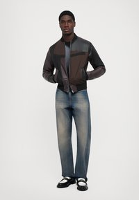 Man i en patchwork bomberjacka av svart och brunt läder, urtvättade blå jeans och svarta och vita loafers mot en vit bakgrund.