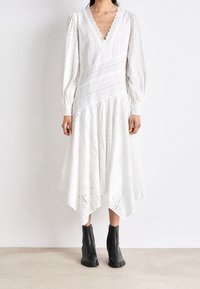 Abito bianco al ginocchio con maniche a sbuffo, scollatura scallop, motivo ad occhiello e orlo asimmetrico. Indossato con un paio di stivaletti neri alla caviglia.