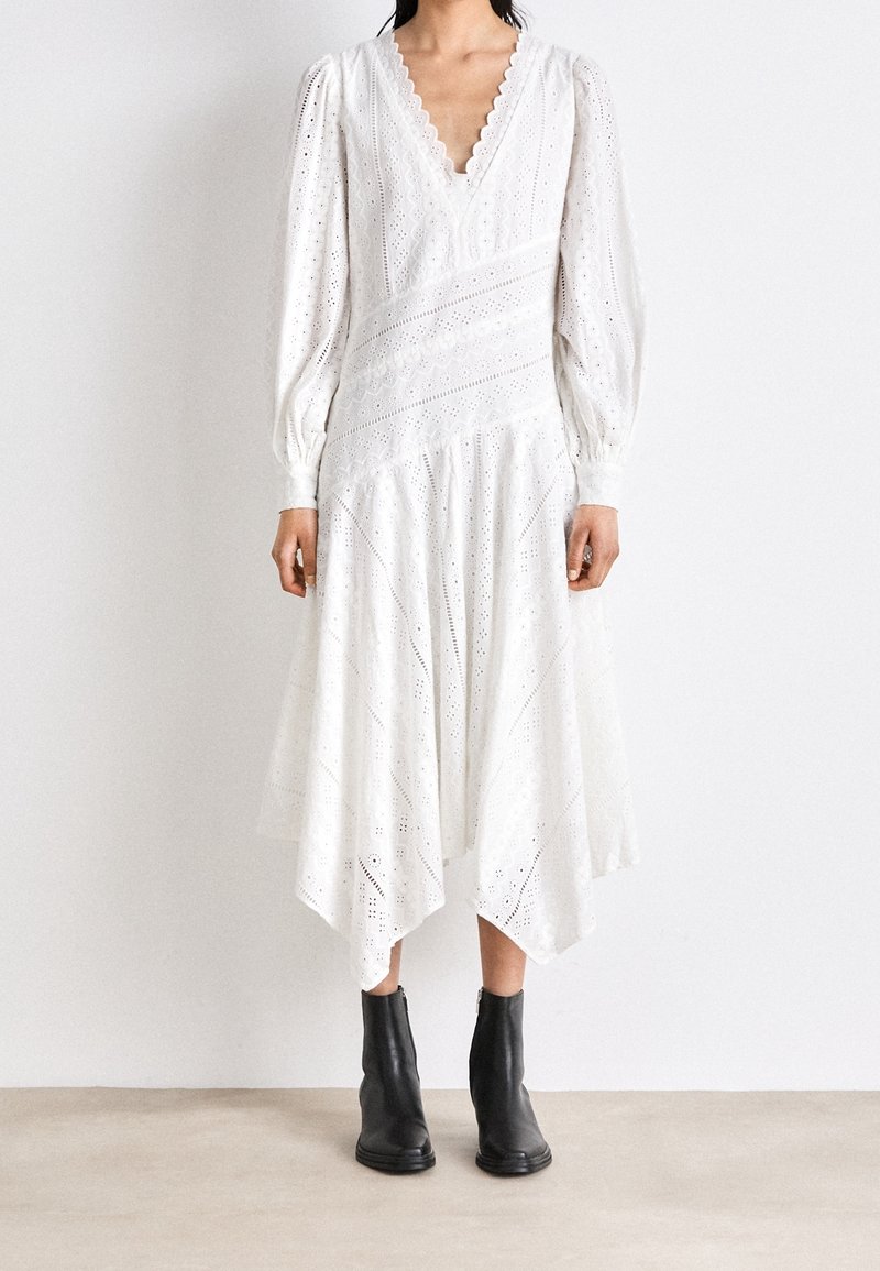 Abito bianco al ginocchio con maniche a sbuffo, scollatura scallop, motivo ad occhiello e orlo asimmetrico. Indossato con un paio di stivaletti neri alla caviglia.