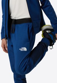 Modre trenirke s črnim pasom in logotipom; gladka tkanina; opazni šivi in poudarki na spodnjih delih nog; kombinirane s črno-modrimi čevlji.