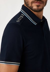 Marineblauwe polo met een klassieke snit, voorzien van een geribde kraag met lichtblauwe accenten en zijborduring van de merknaam "GUESS."