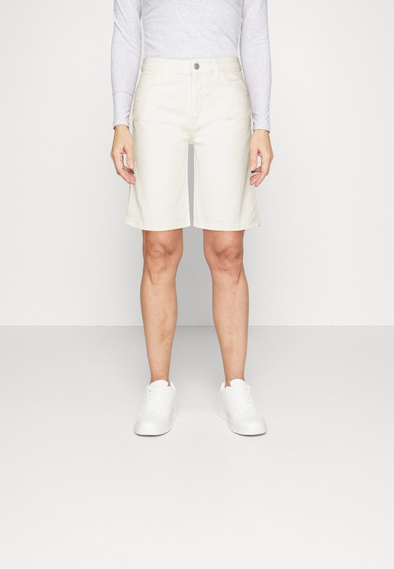 Pantalones cortos beige de corte recto, hechos de algodón, con cierre de botón en la parte delantera y bolsillos laterales. Combinados con zapatillas blancas.