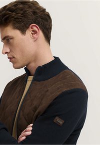 Kurtka zapinana na zamek w kolorze granatowym z brązowym pikowanym przodem. Posiada wysoki kołnierz oraz teksturowany materiał, a na rękawie znajduje się mała metka z logo.
