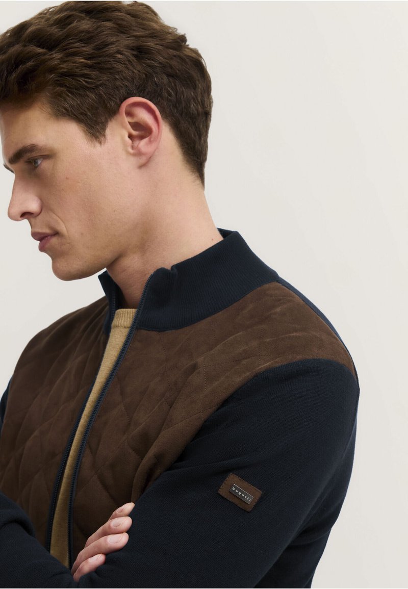 Kurtka zapinana na zamek w kolorze granatowym z brązowym pikowanym przodem. Posiada wysoki kołnierz oraz teksturowany materiał, a na rękawie znajduje się mała metka z logo.