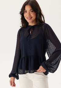 Blusa lungo in un elegante blu navy, con maniche lunghe, collo rotondo, dettaglio a fiocco, chiusura con bottoni, tessuto testurizzato e vita a peplo.