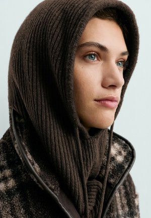 Jeune personne aux yeux bleus portant une capuche en tricot côtelé marron et une veste polaire à motifs, regardant légèrement vers le haut et vers la droite.