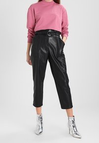 Mujer que lleva una camiseta de manga larga rosa, pantalones de cuero negros de tiro alto y ajustados con cinturón, y botas de tacón alto plateadas hasta el tobillo.