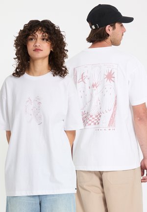 Jonge vrouw die naar voren kijkt en man met de rug naar voren, beiden dragen witte T-shirts met abstracte rode grafische ontwerpen en casual broeken tegen een effen achtergrond.