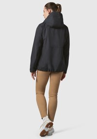 Črna vodoodporna jakna s kapuco, podrobnosti o elastičnem manšetnem delu in sproščen fit. Nosi se nad svetlo rjavimi ozkimi hlačami in belimi športnimi copati.