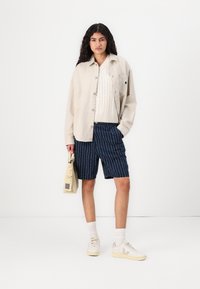 Beige skjorte med knapper over en hvit strikket genser, marineblå shorts med hvite vertikale striper, kremfargede joggesko, og en beige bag.