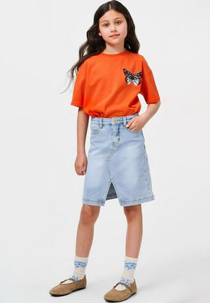 Dziewczyna z długimi ciemnymi włosami w pomarańczowej koszulce z nadrukiem motyla, jasnej dżinsowej spódnicy, wzorzystych skarpetkach i brązowych butach, stojąca z ręką na biodrze.