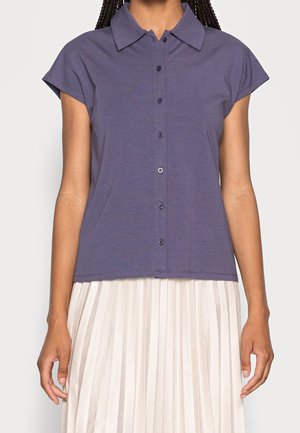 Person wearing a short-sleeve, button-up purple shirt with a wide collar and a white pleated skirt, standing against a plain background.