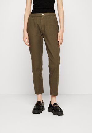 Femme portant un pantalon fuselé vert olive avec des poches plaquées à l'avant, associé à des mocassins à plateforme noirs et un haut sans manches noir.