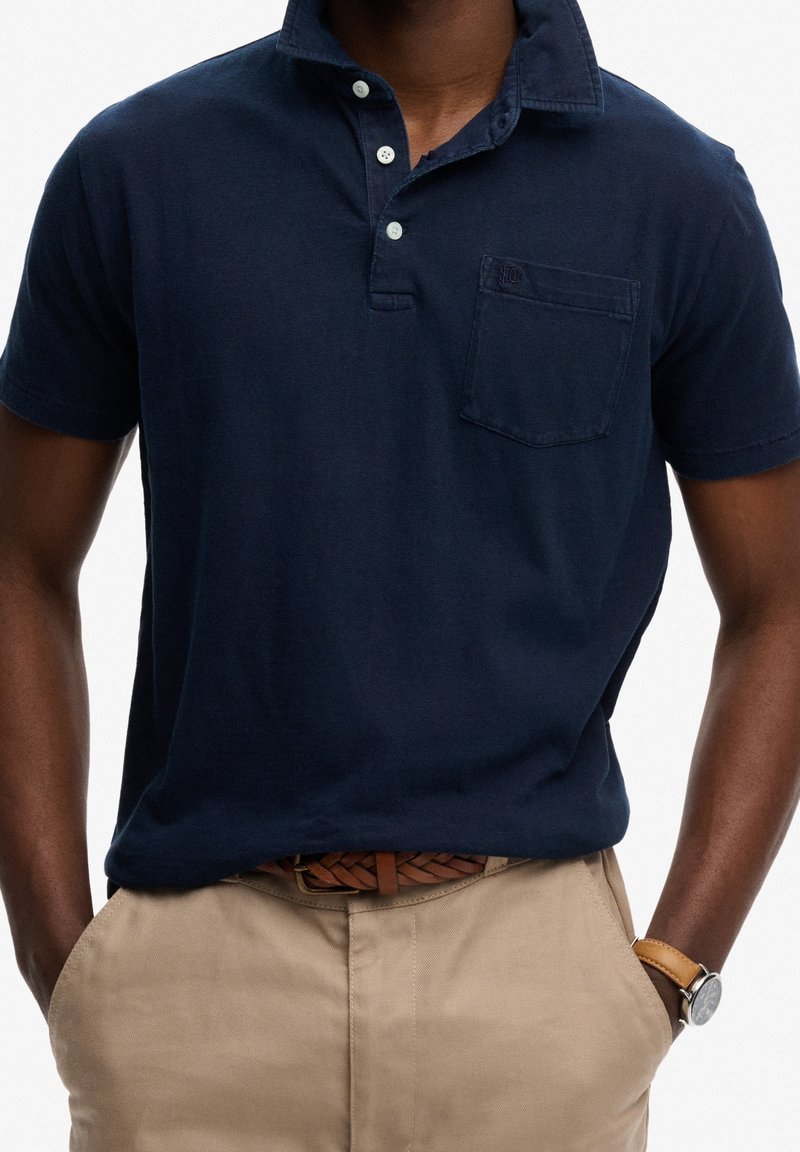 Homme portant un polo bleu marine avec poche poitrine, pantalon beige, ceinture tressée marron et montre-bracelet avec bracelet en cuir marron, mains dans les poches.