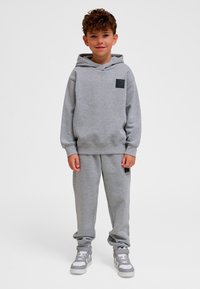 Grå sweatshirt med huva och framficka, matchad med grå mjukisbyxor. Har en svart logotypmärke på bröstet och fickan.