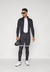 Radbekleidung mit einer schwarzen, taillierten Jacke, einem weißen Jersey, schwarzen gepolsterten Shorts mit weißen Streifen und weißen Radschuhen. Helm in der Hand.